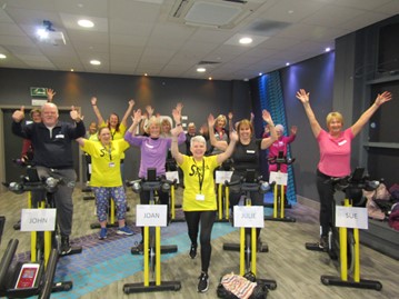 Group of older people, in bright yellow 'senior spin' tops, in the studio with static bikes
