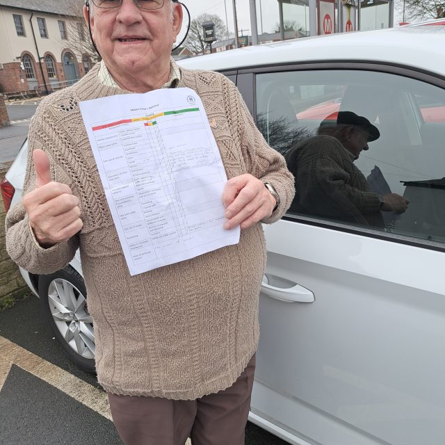 Older man, wearing a cap, standing next to a caar and holding up a piece of paper - his appraisal.