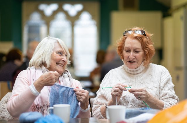 Two women smiling and knitting with coffees