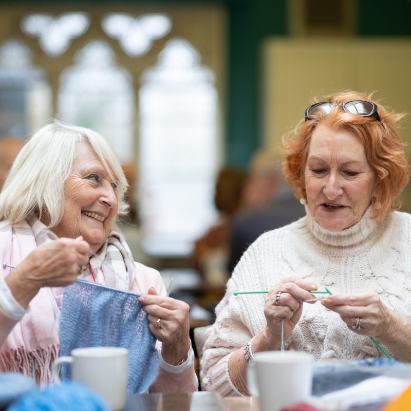 Two women smiling and knitting with coffees