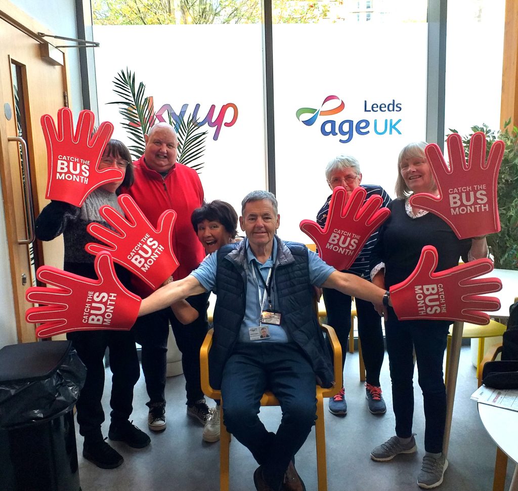 Group of 7 people, each wearing one (or two) huge red 'hands', each of which has the text "CATCH THE BUS MONTH" across the 'palm'.. In the background are windows, with "skyup" and "AgeUK Leeds" showing on the glass.