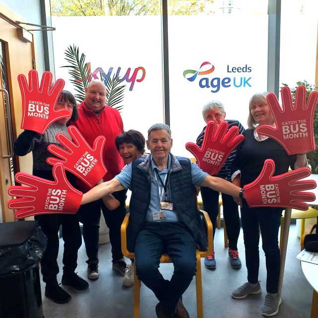 Group of 7 people, each wearing one (or two) huge red 'hands', each of which has the text "CATCH THE BUS MONTH" across the 'palm'.. In the background are windows, with "skyup" and "AgeUK Leeds" showing on the glass.