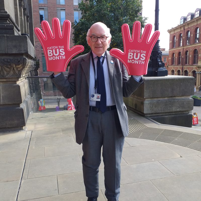 Older man standing outside, wearing a huge red 'hand' on each hand,, on which the text "CATCH THE BUS MONTH"